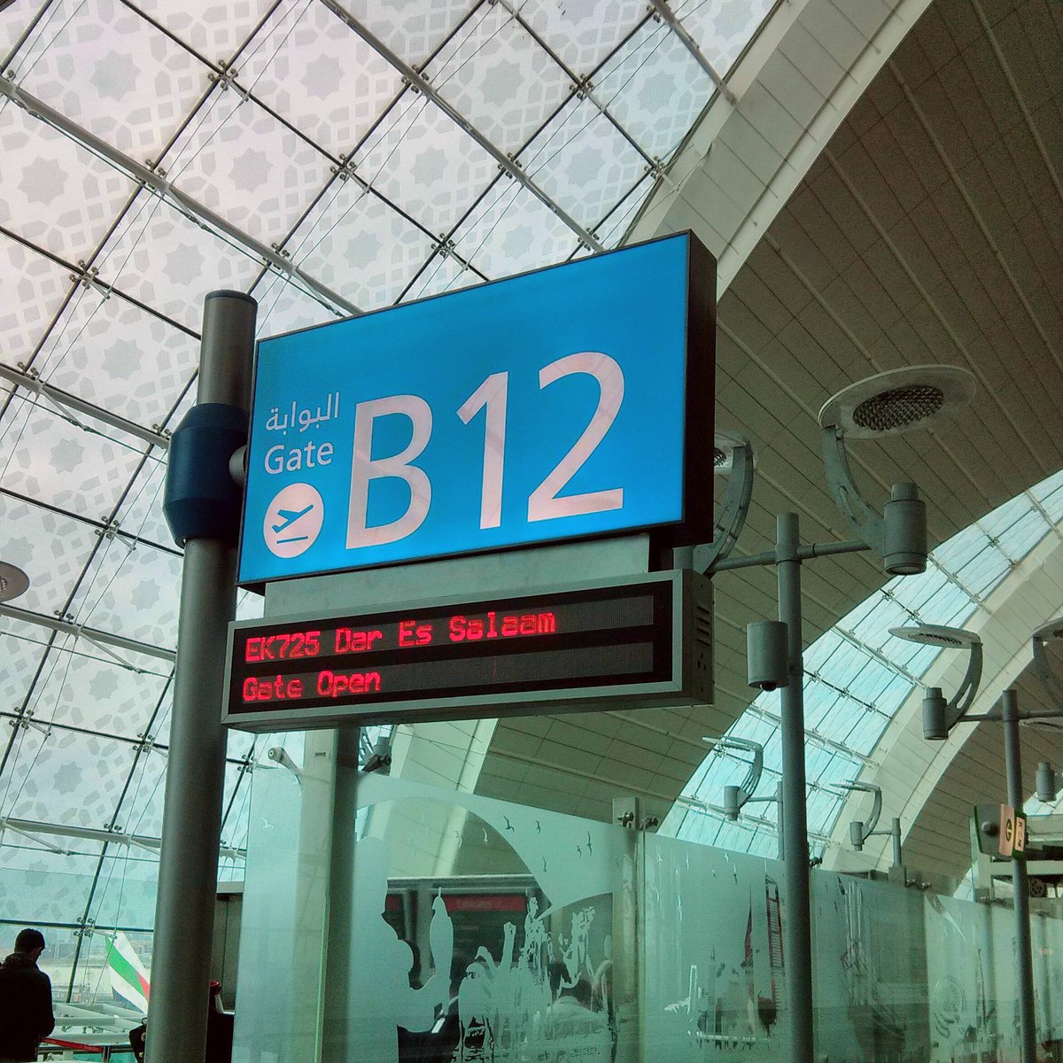 Ein Schild am Flughafen Dubai (DXB), das in Weiß auf Himmelblau das Gate B12 anzeigt. Eine digitale Anzeige darunter weist auf den Flug EK725 nach Daressalam hin.