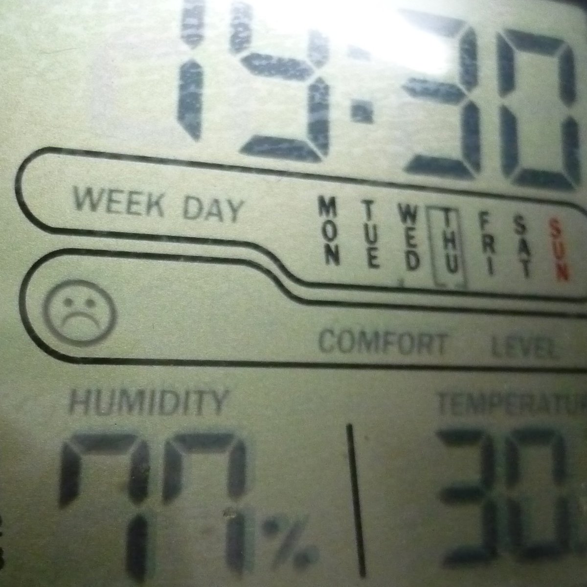 Die digitale Anzeige einer Klimaanlage zeigt 30°C Temperatur und 77% Luftfeuchtigkeit an. Ein Emoticon mit negativem Gesichtsausdruck verdeutlicht die unangenehme Wetterlage.