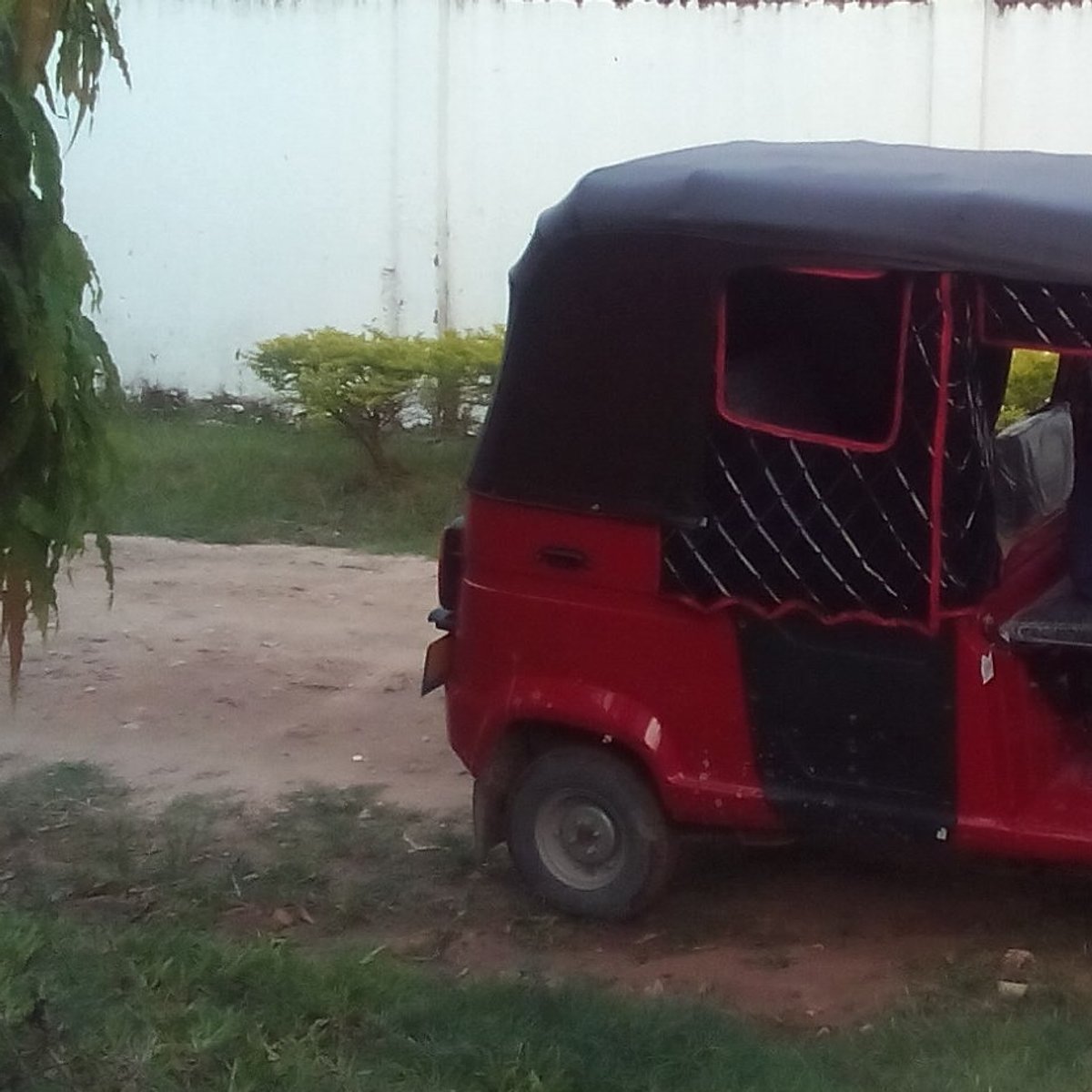 Ein rotes Bajaj (auch Bajaji oder Autorikscha) mit schwarzem Dach. Das Bajaj steht auf einem Sandweg vor einer weißen Mauer. Ein Bajaj ist ein motorisiertes Fahrzeug mit drei Rädern.