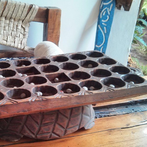 Ein Bao-Spielbrett aus Holz, das aus einer Holzplatte mit 32 Mulden besteht. In den Mulden befinden sich je zwei Pflanzensamen, die als Spielsteine verwendet werden.
